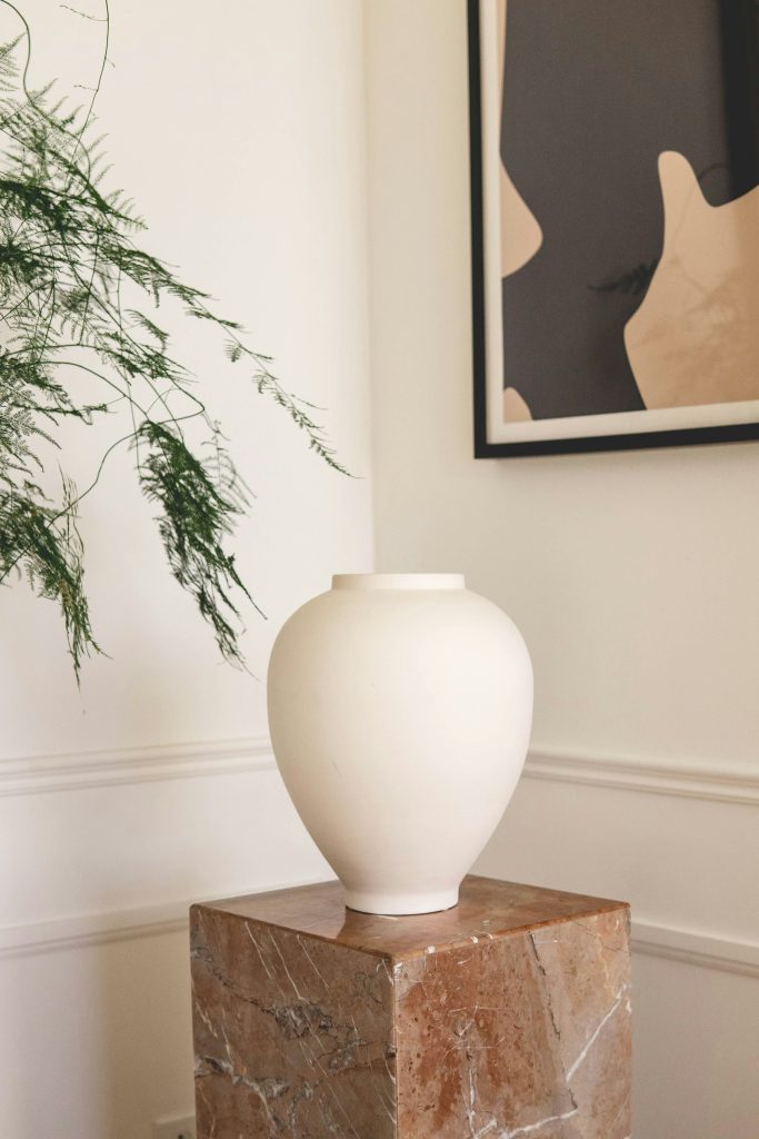 One single white oval vase that is thinner towards the bottom, sitting on top a light brown rectangle chunk of marble in the corner of a white room